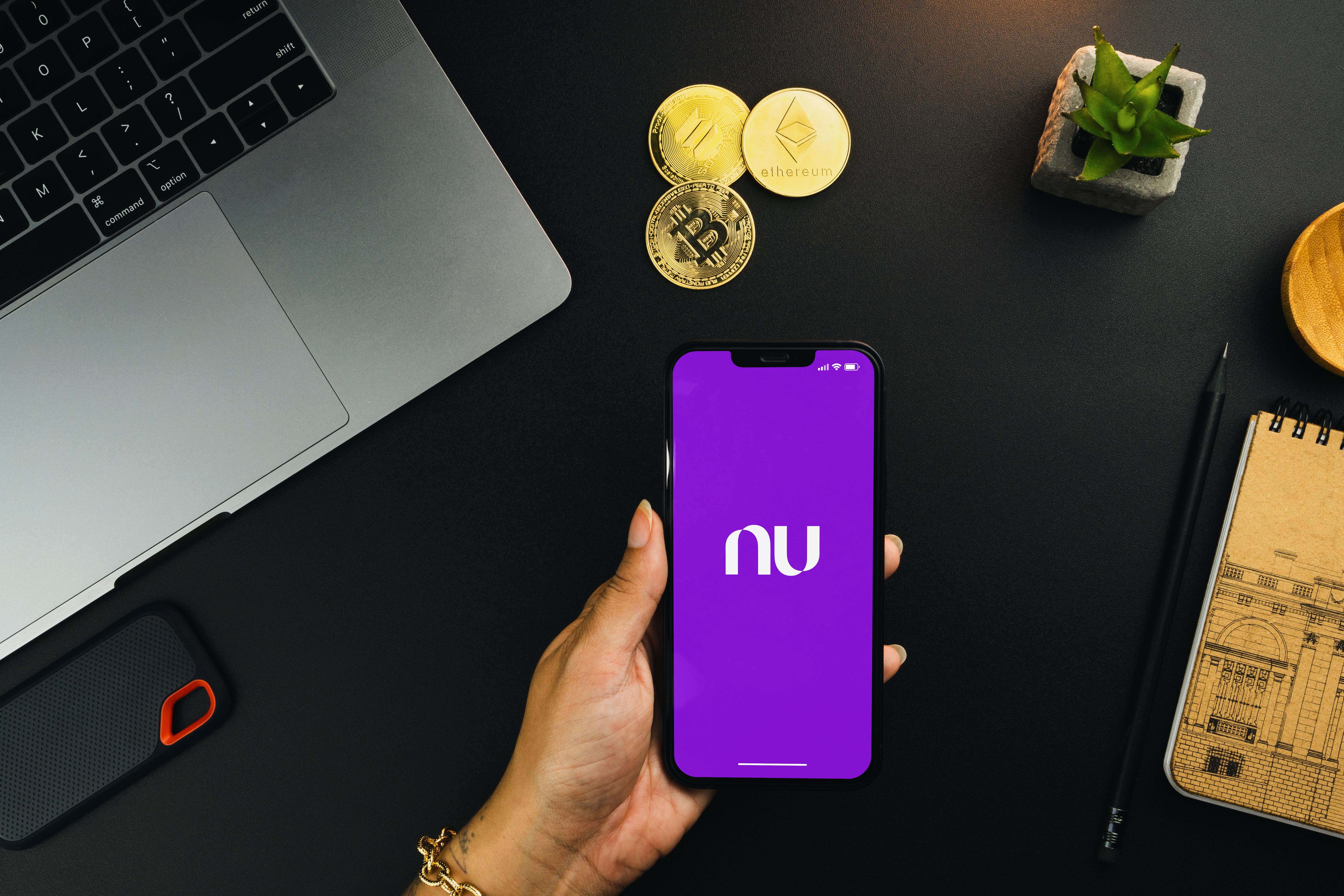 A person holds a smartphone with the Nubank bank app running on the screen. On a table behind the phone, artistic representations of cryptoassets are visible, along with a laptop computer.