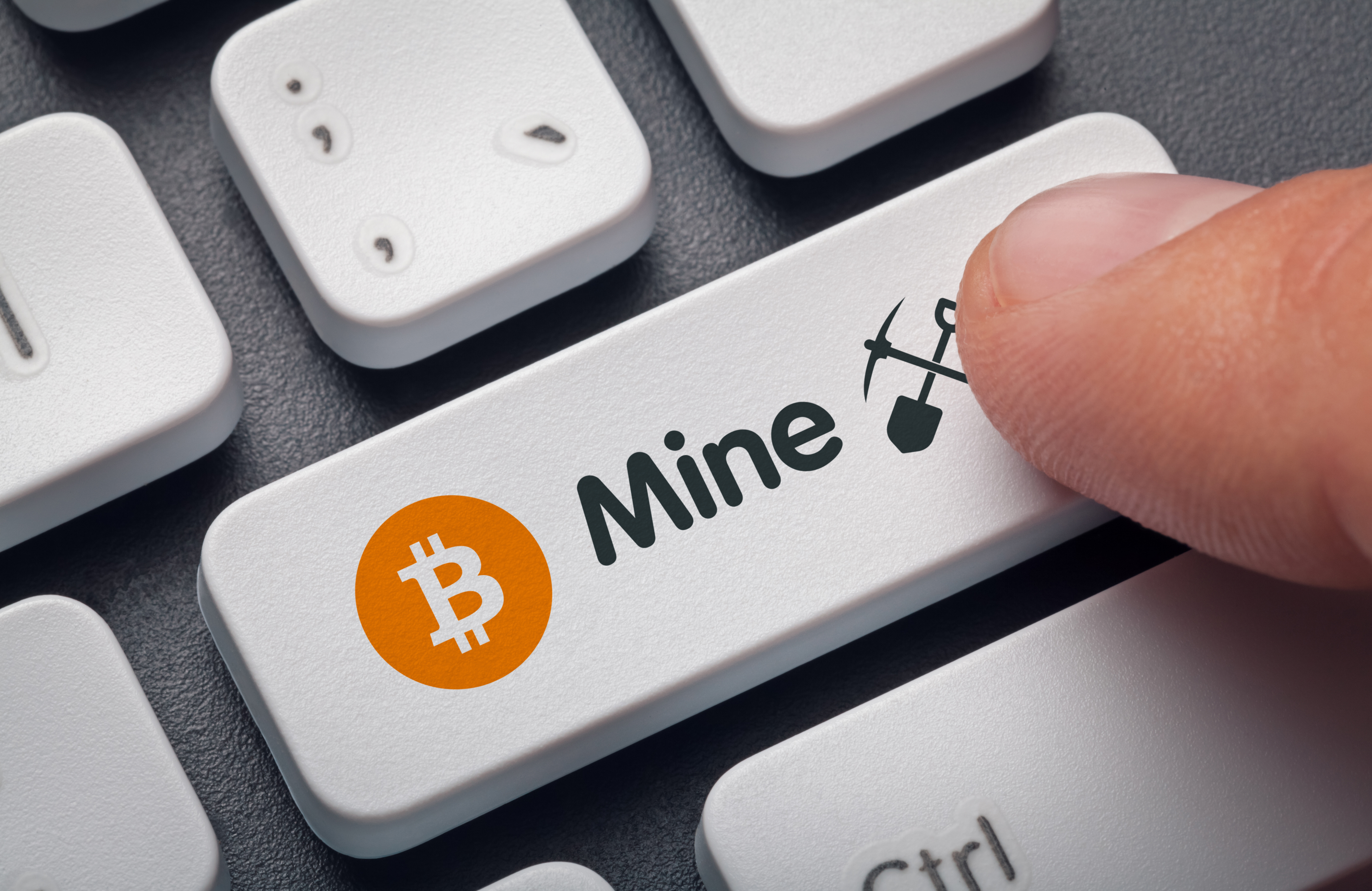 A person&amp;rsquo;s finger presses a computer keyboard key. The key is decorated with a symbol that represents Bitcoin, as well as the word &amp;ldquo;mine&amp;rdquo; and drawings of a pick and shovel.