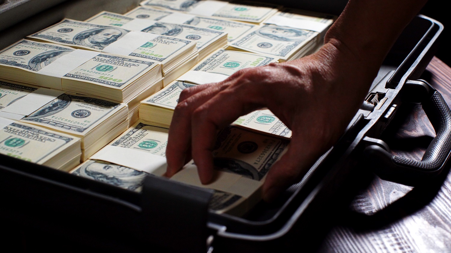 A person&amp;rsquo;s hand grabs a pile of US dollar bills from a suitcase filled with bundles of cash.