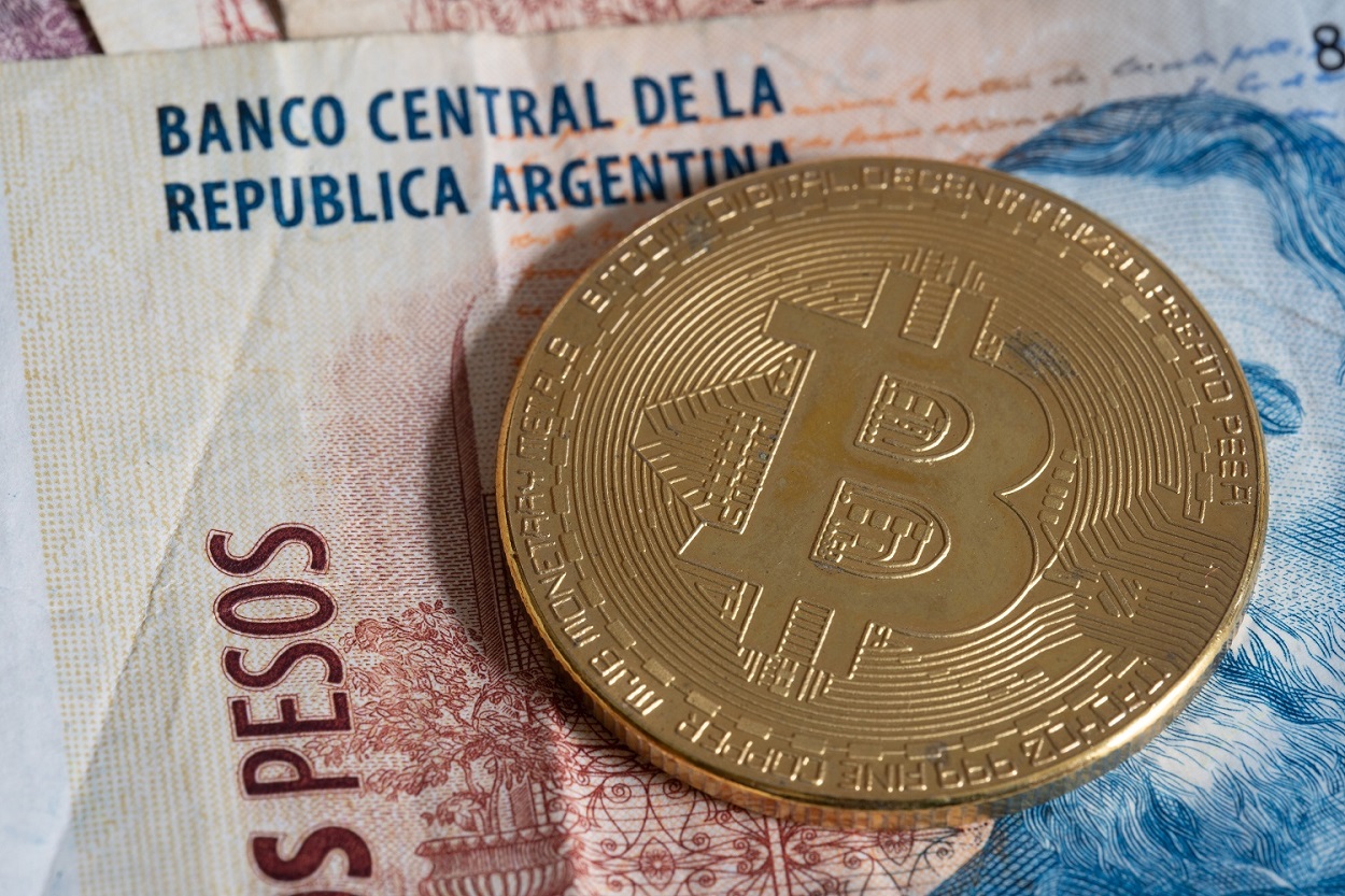 A metal coin represnting Bitcoin rests on top of an Argentine banknote.