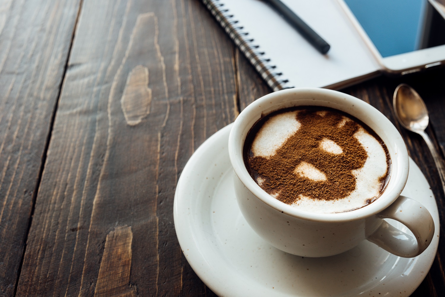 A cup of coffee on a wooden table next to a notepad and a mobile phone, decorated with cinnamon powder made to resemble the Bitcoin logo.
