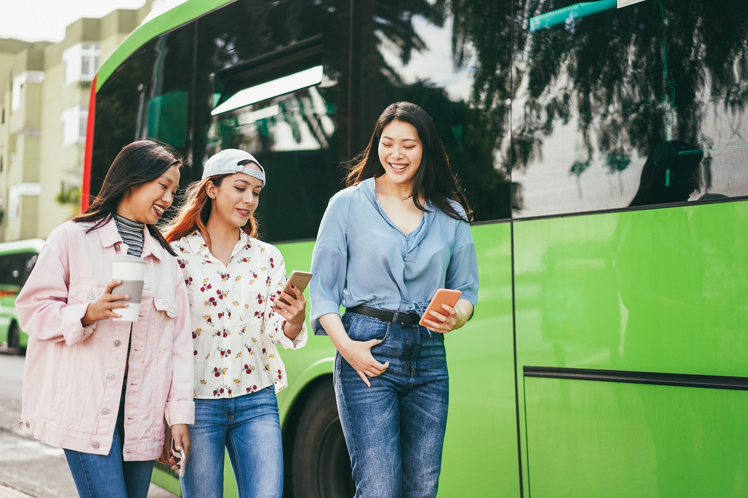 Three young women look at their mobile phones as they walk past a bus.