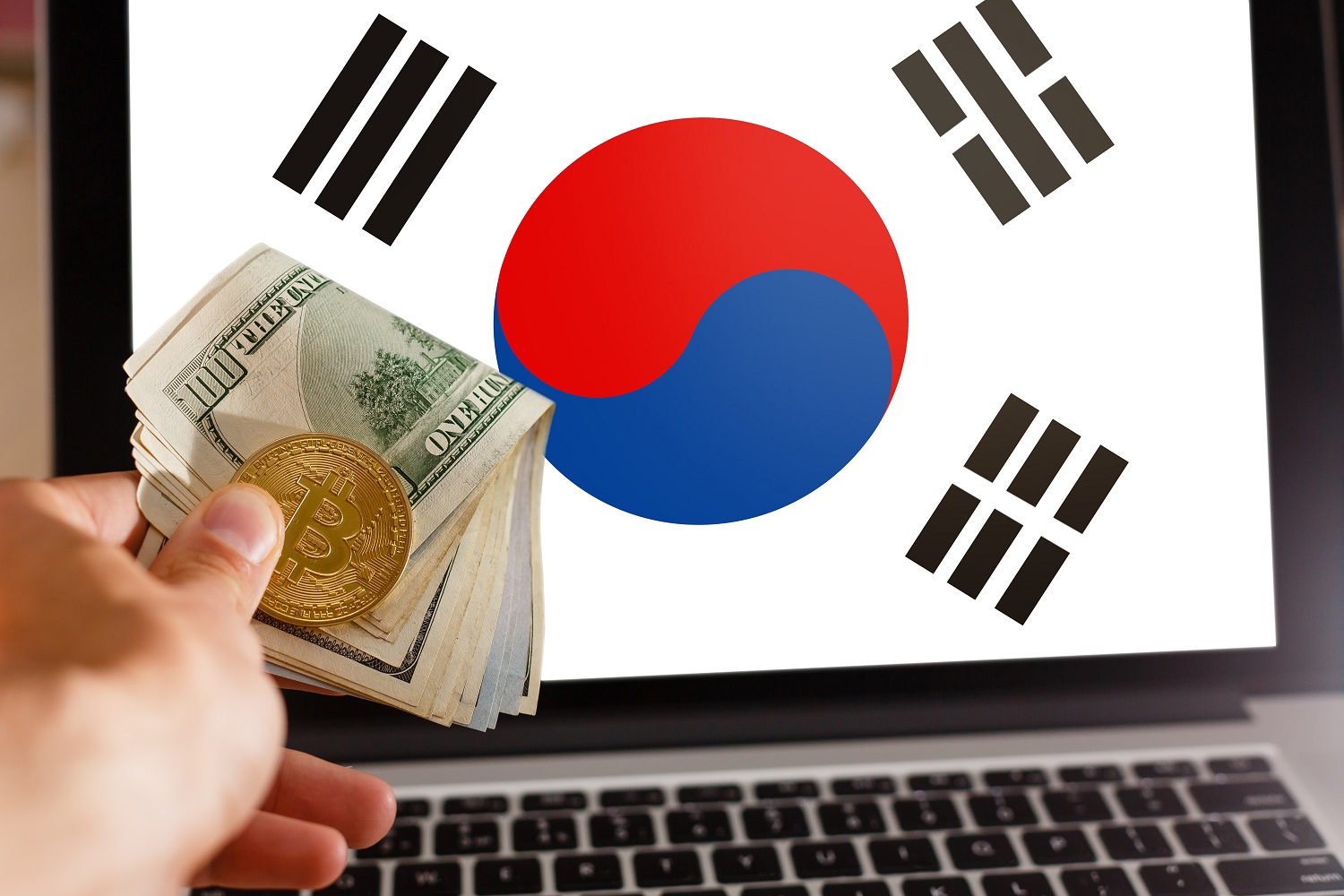 A hand holds US dollar banknotes, as well as a metal token intended to represent Bitcoin in front of a laptop screen with a South Korean flag displayed on its screen.