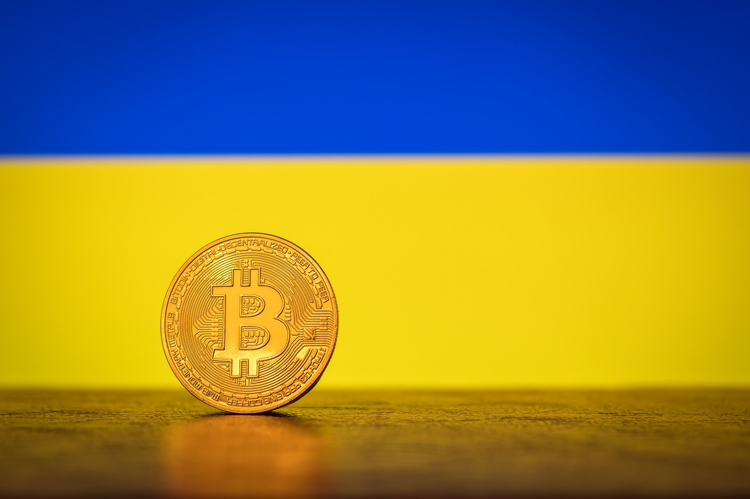 A metal coin intended to represent Bitcoin on a wooden table with their Ukrainian flag in the background.