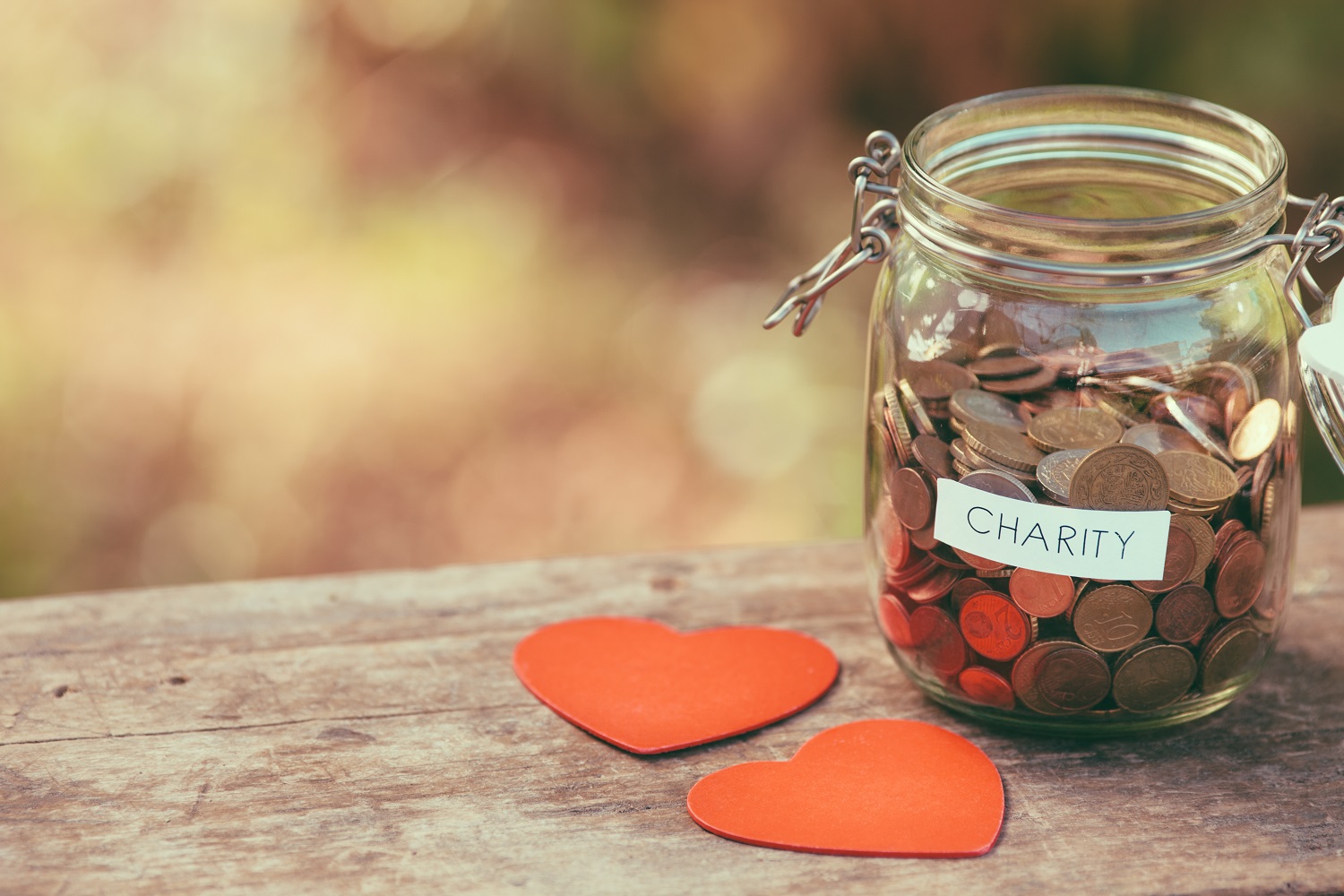 A jar of coins with the word “Charity” written on a label on the side rests on a table. Two red paper hearts are next to the jar.