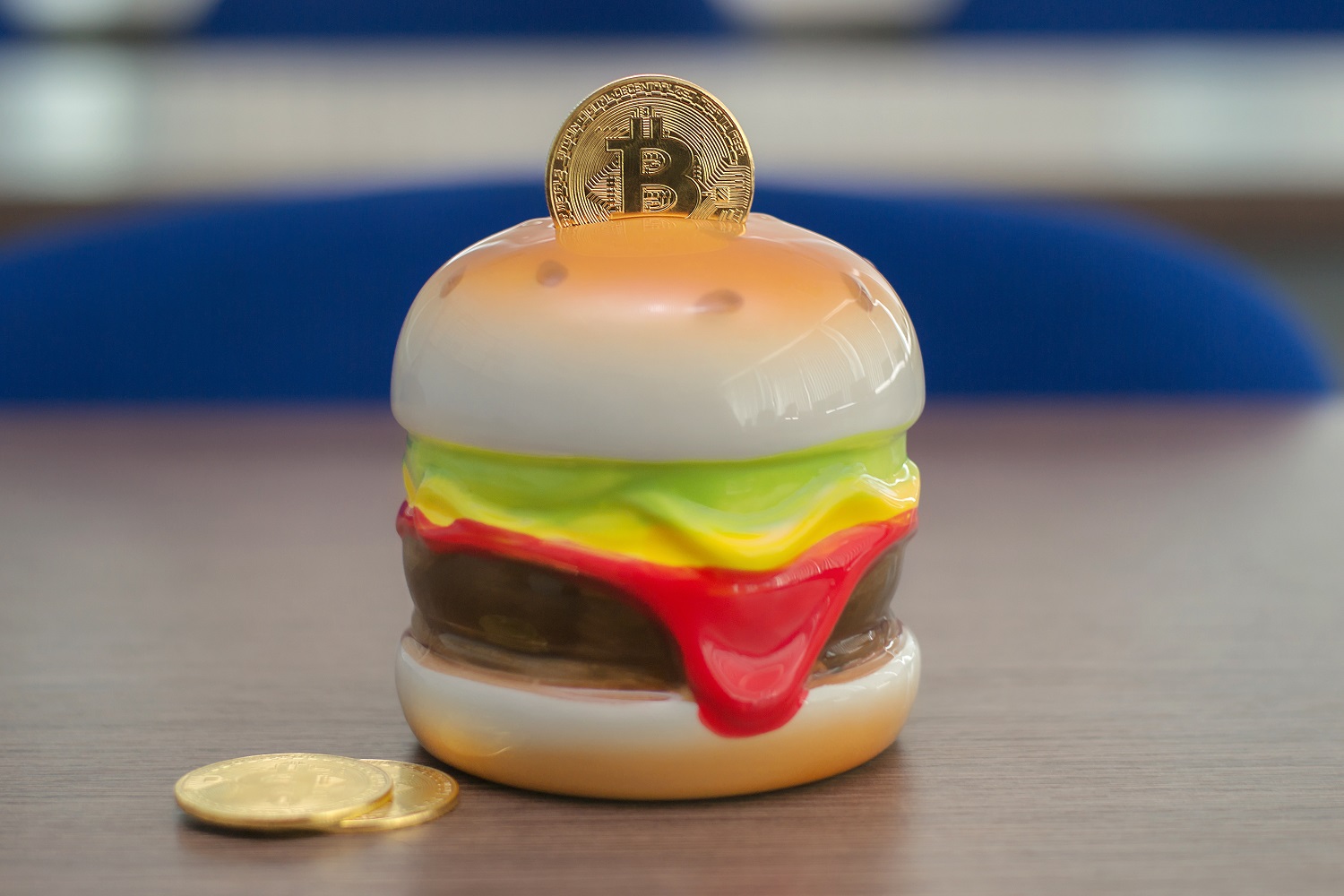 A metal coin intended to represent Bitcoin in the slot of a piggy bank made to look like a burger on a table next to other metal tokens.
