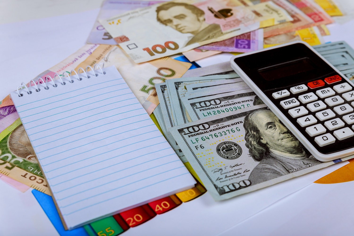 A calculator and a paper notepad rest atop a selection of USD and UAH banknotes.