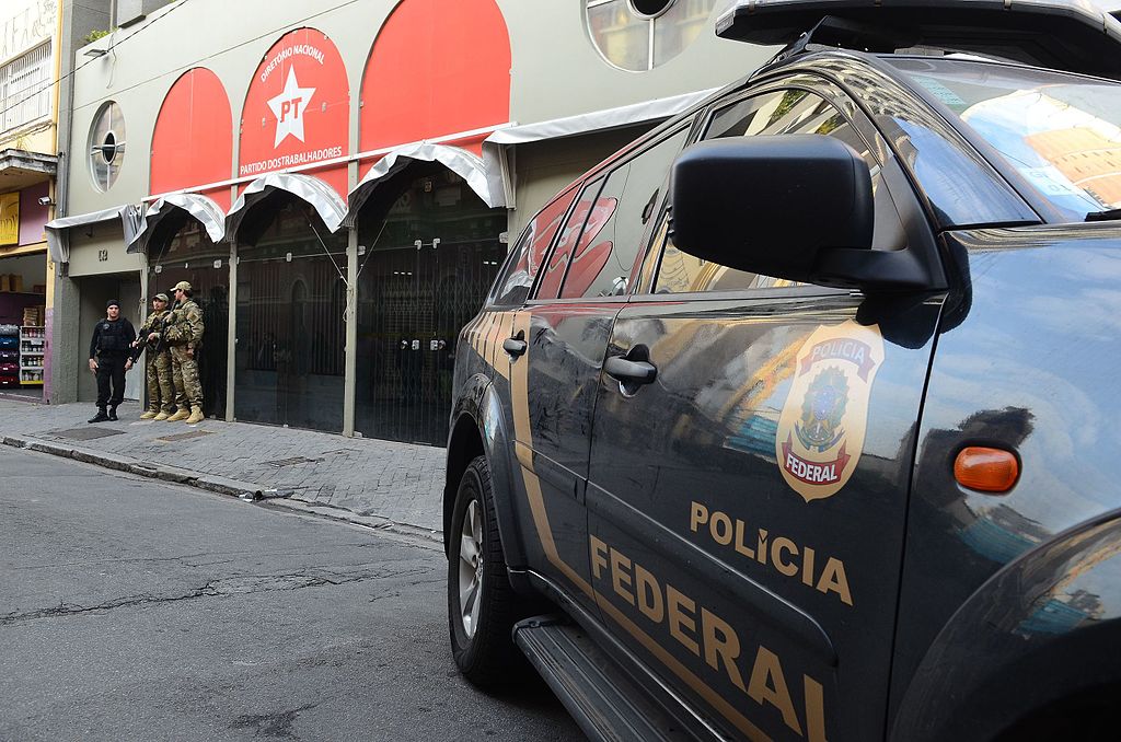 Armed Brazilian police and army officers stand outside a building with a police vehicle in the foreground.