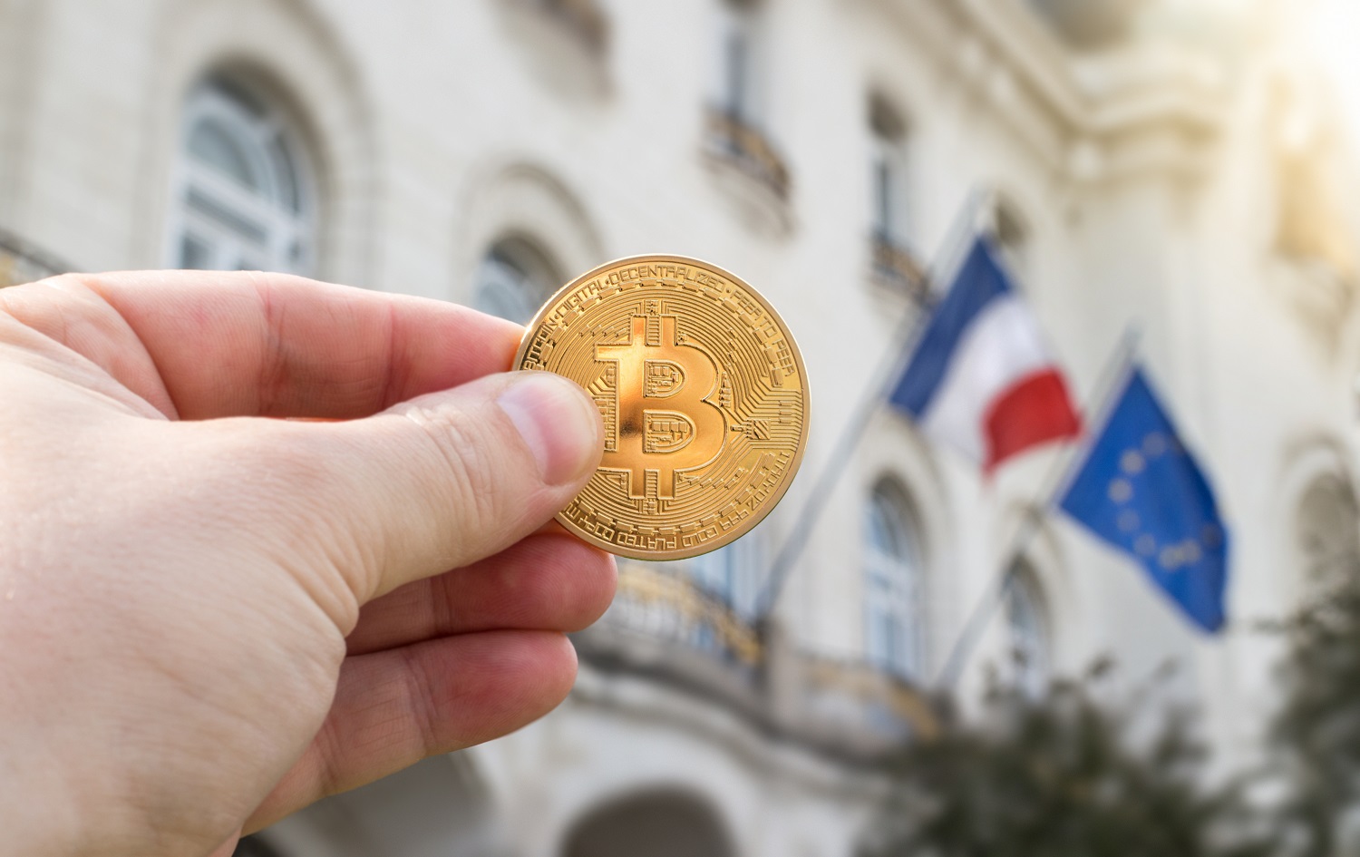 A person’s hand holds up a metal token intended to represent Bitcoin against the background of a building and the EU and French flags.