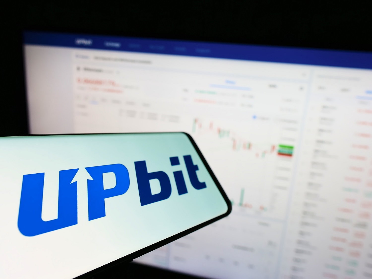 A mobile phone displaying the logo of the South Korean crypto exchange Upbit against the background of a laptop monitor showing an price graph.