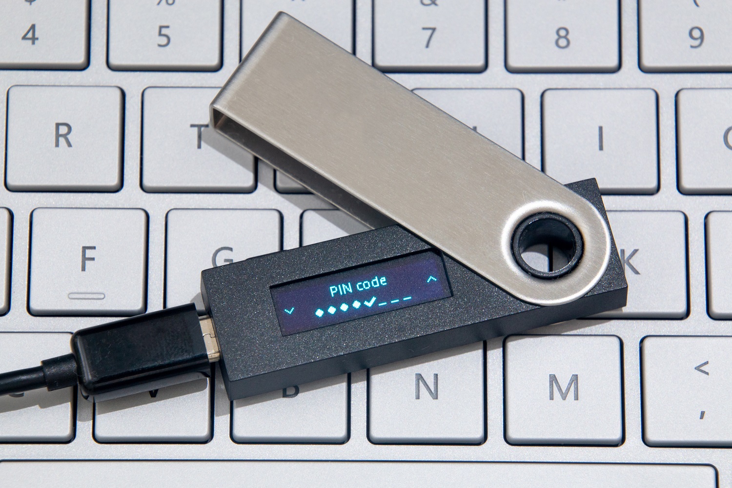 A crypto hardware wallet, plugged in to a cord via a USB socket, rests on a laptop keyboard.