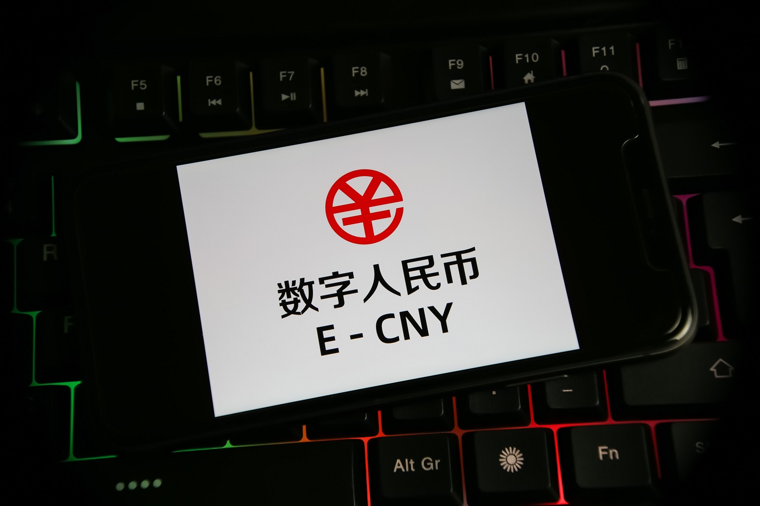 A closeup view of a mobile phone screen resting on a computer keyboard with the logo and symbol of the Chinese digital currency, the e-CNY.