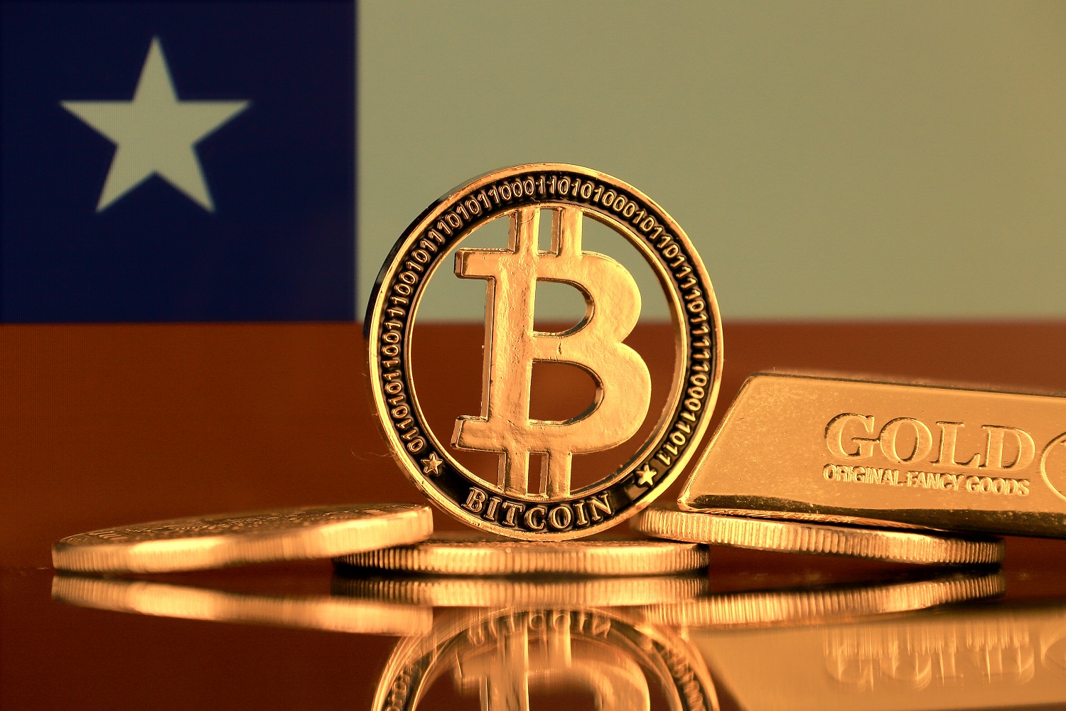 A metal token intended to represent Bitcoin, next to other tokens, a gold bar, against the backdrop of the Chilean Flag.