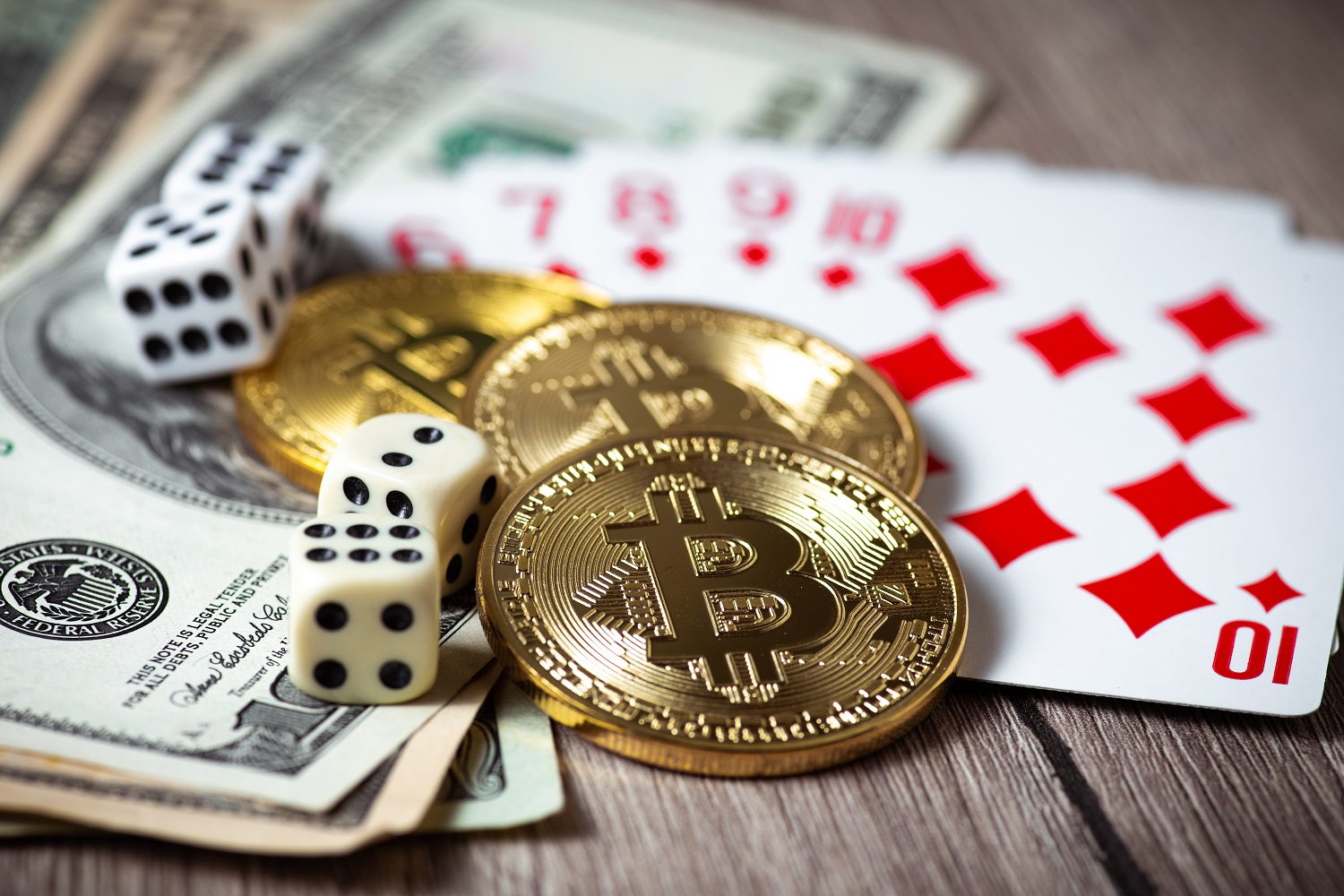 Three tokens intended to represent Bitcoin, along with playing cards, dice, and banknotes, on a wooden tabletop.