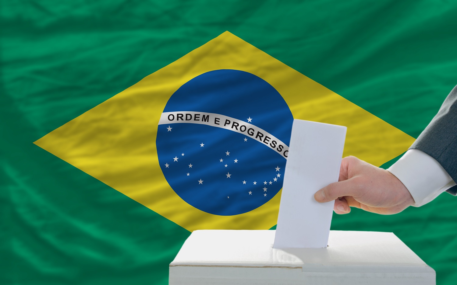 A man places a voting slip into a ballot box against the backdrop of the Brazilian flag.