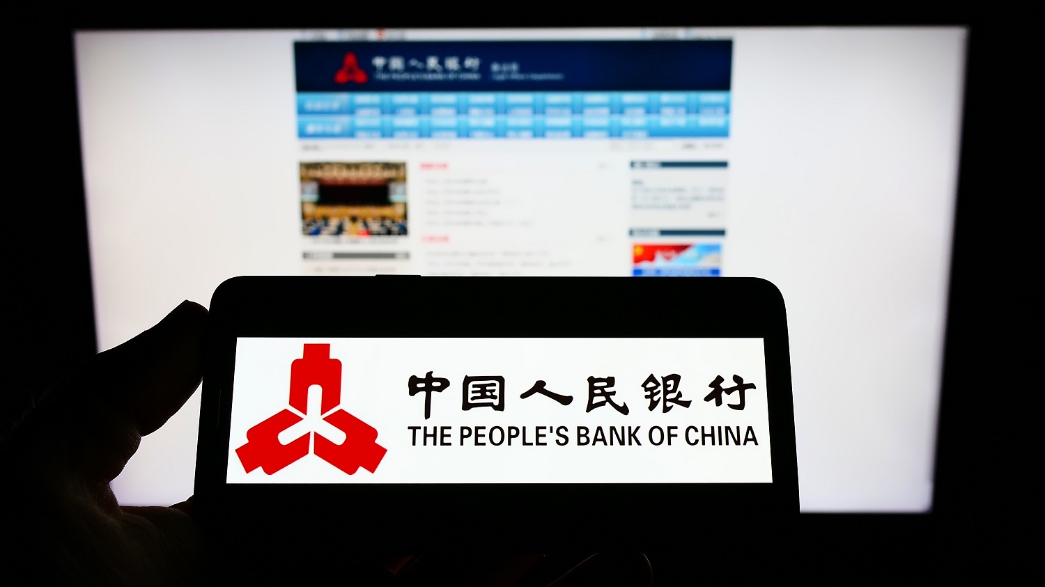 A person holds a cellphone with the logo of the People’s Bank of China on its screen in front of a PC displaying the bank’s webpage.