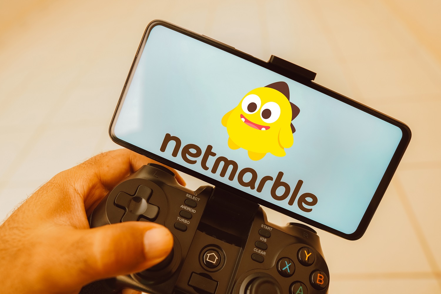 A person holds a joystick attached to a mobile phone with the Netmarble logo displayed on the phone’s screen.