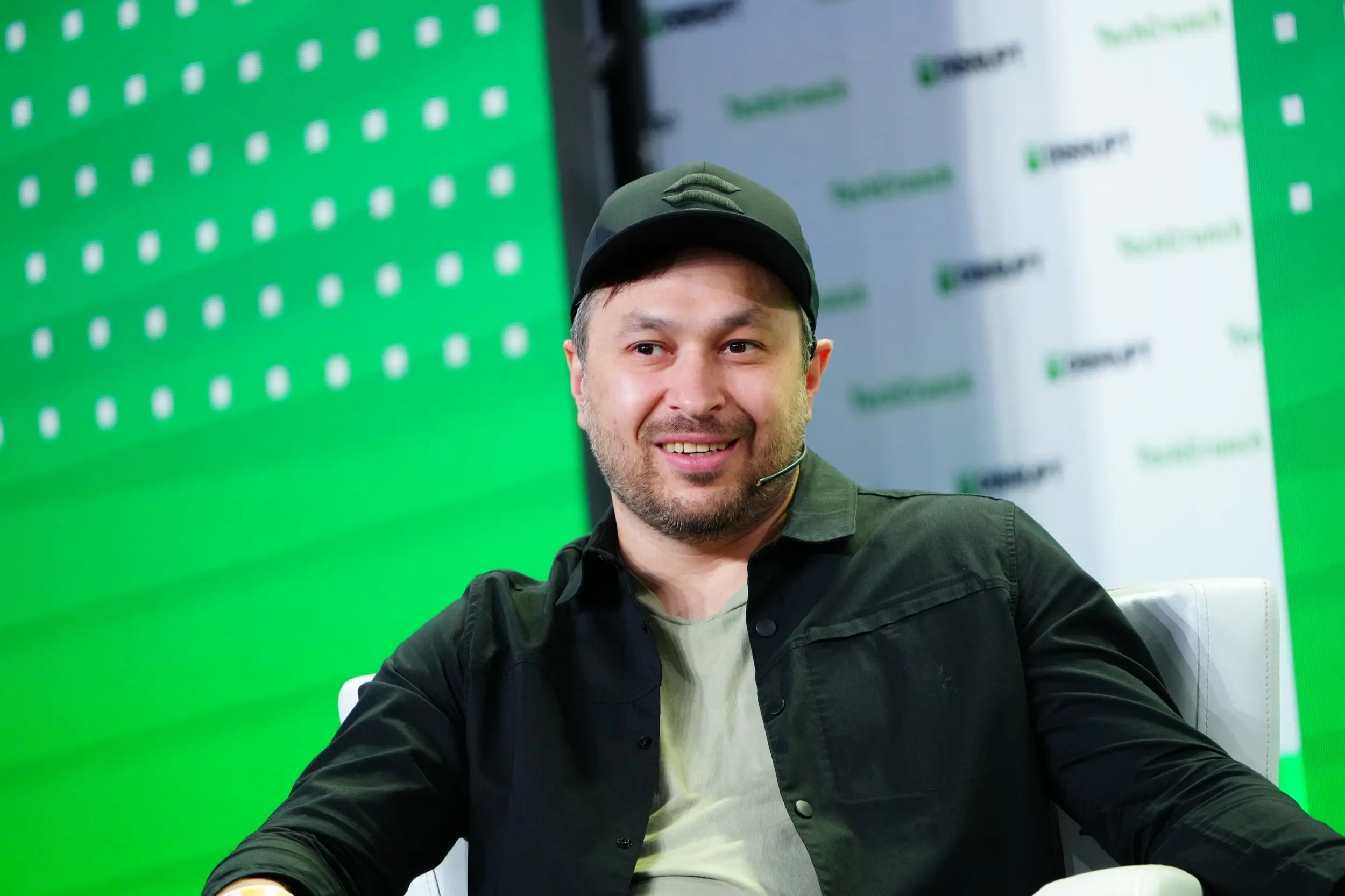 Smiling man in a black shirt and cap sitting on stage during a conference.