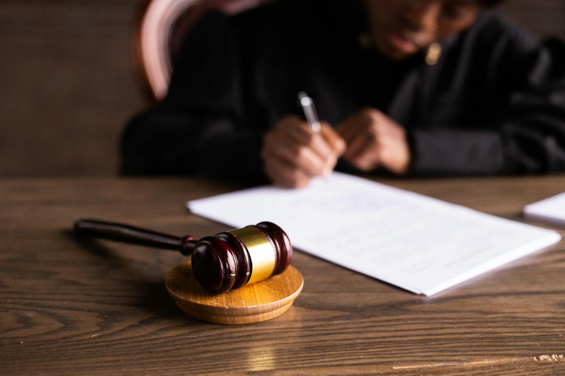 Gavel on a table with a person writing on a document in the background.
