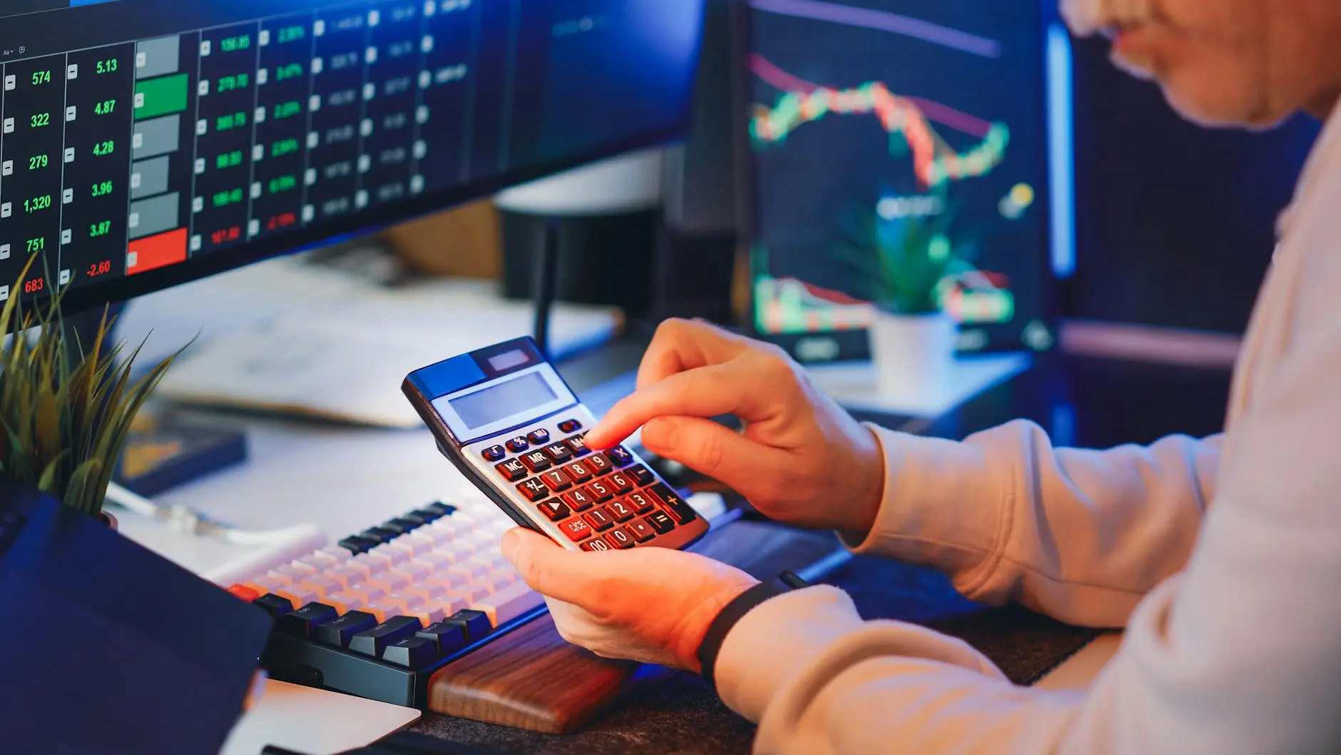 Person using a calculator in front of two screens showing stock market data and charts.