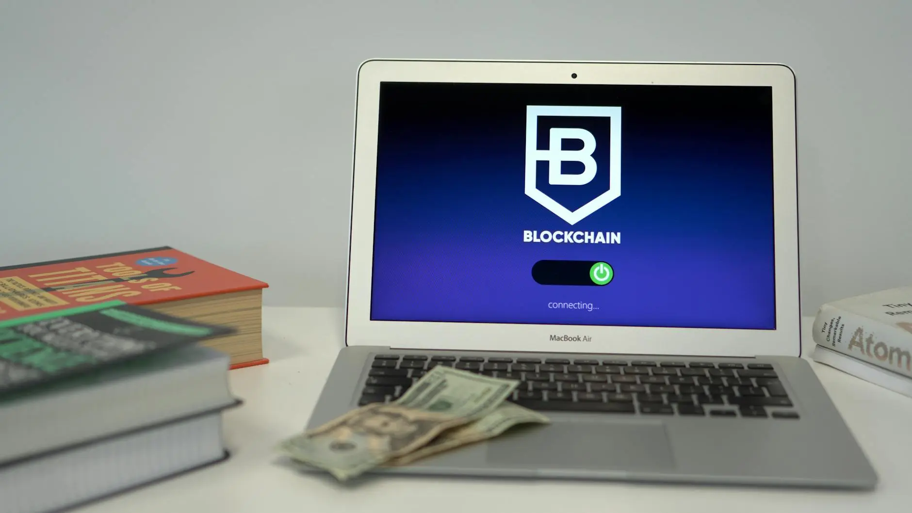 A MacBook Air displaying a blockchain login screen with cash and books nearby.