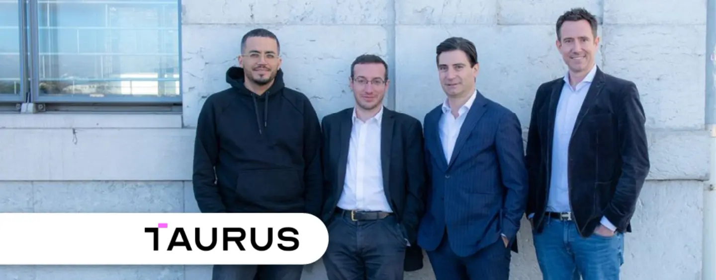 Four individuals standing in front of a stone wall, with a Taurus logo visible.