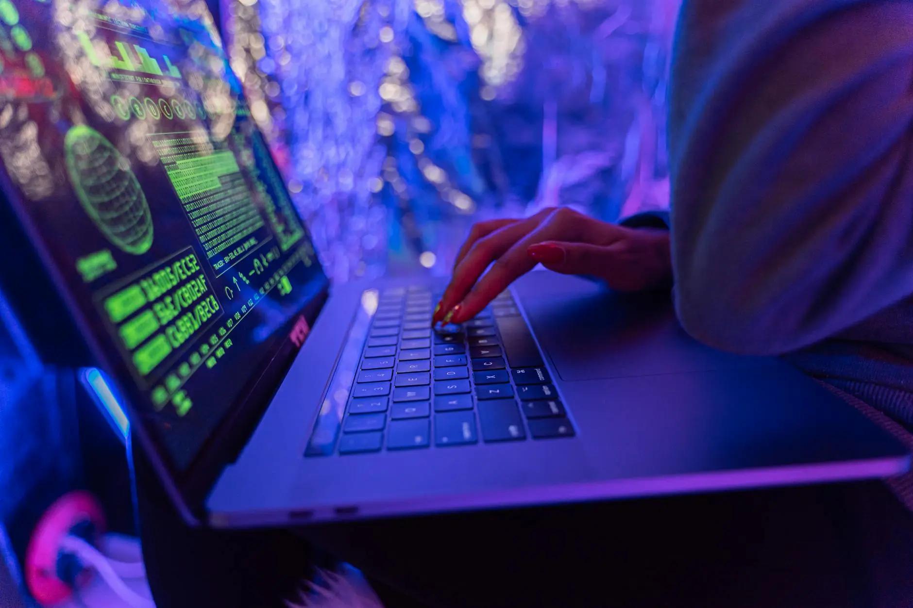 Close-up of hands typing on a laptop with vibrant green and blue code on the screen.