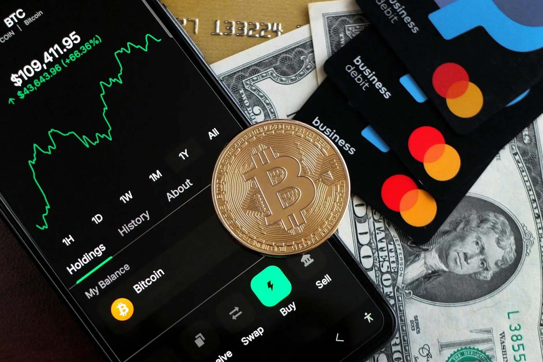 Bitcoin coin on a smartphone displaying trading data, with payment cards and cash in the background.