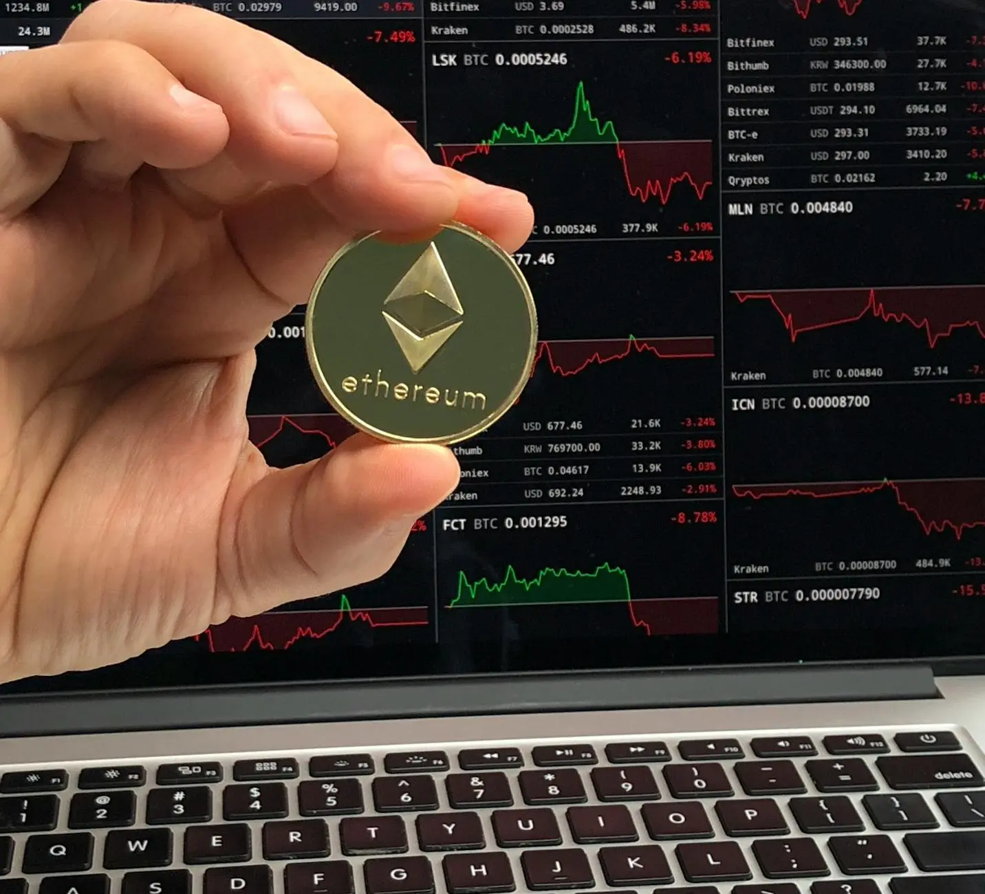 A person holding a gold Ethereum coin in front of cryptocurrency market charts on a laptop.