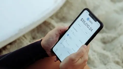 Person holding a smartphone displaying USDC balance of $5,010.24 on a beach.
