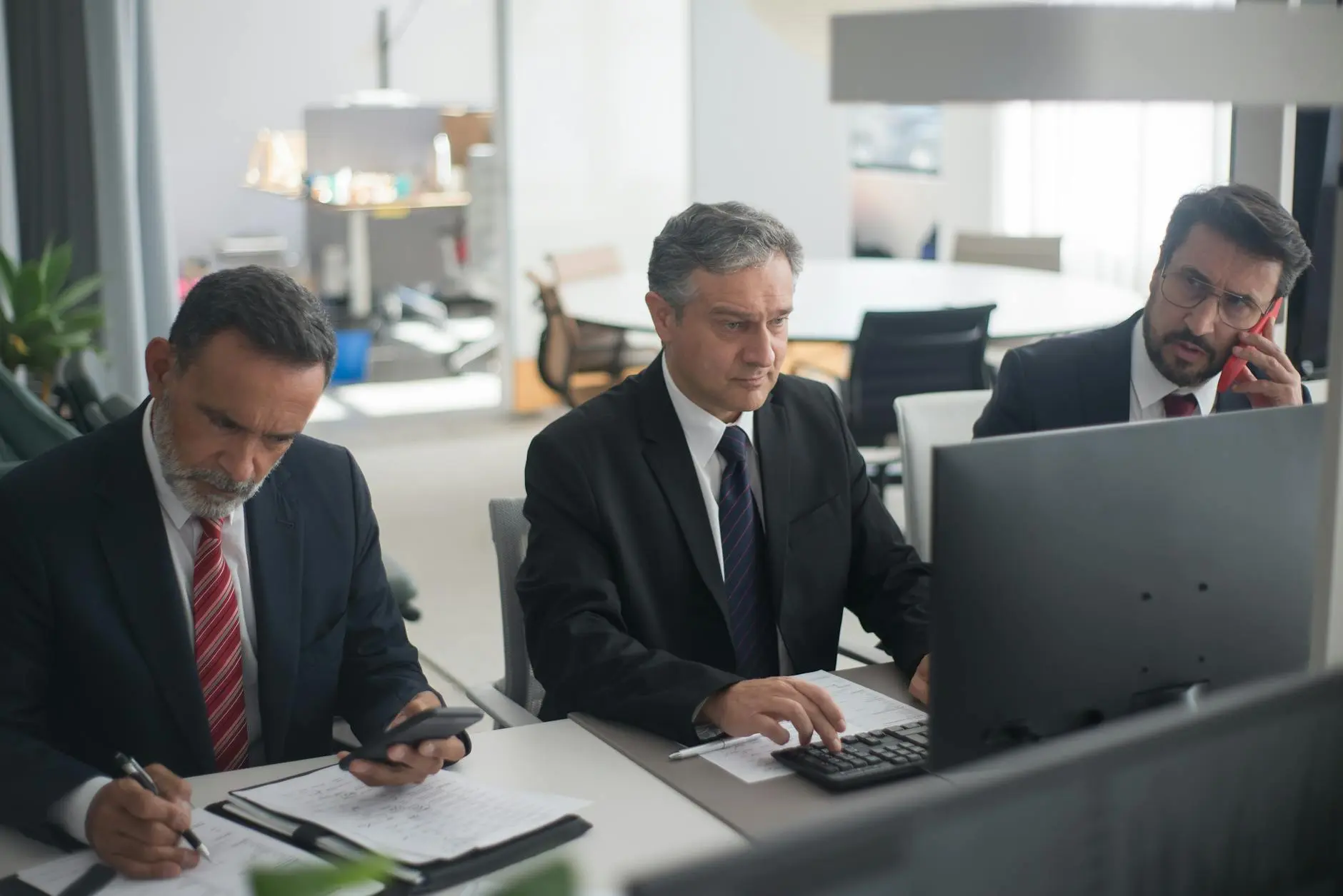 Three men in suits working at a finance office, focused on tasks at their desks.