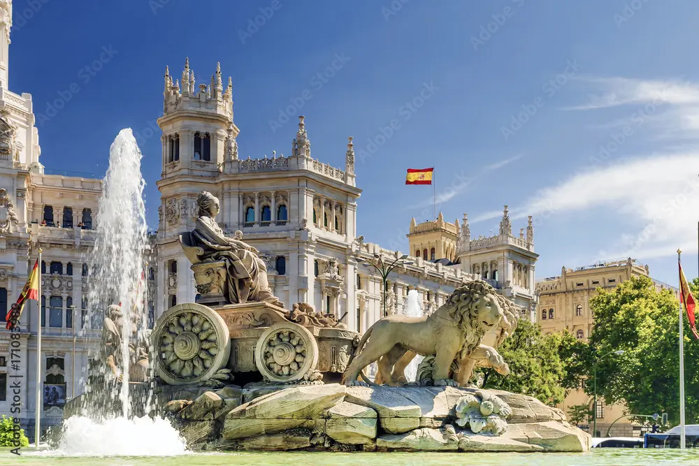 Fountain with statue of a woman and lion, ornate buildings in background against blue sky.