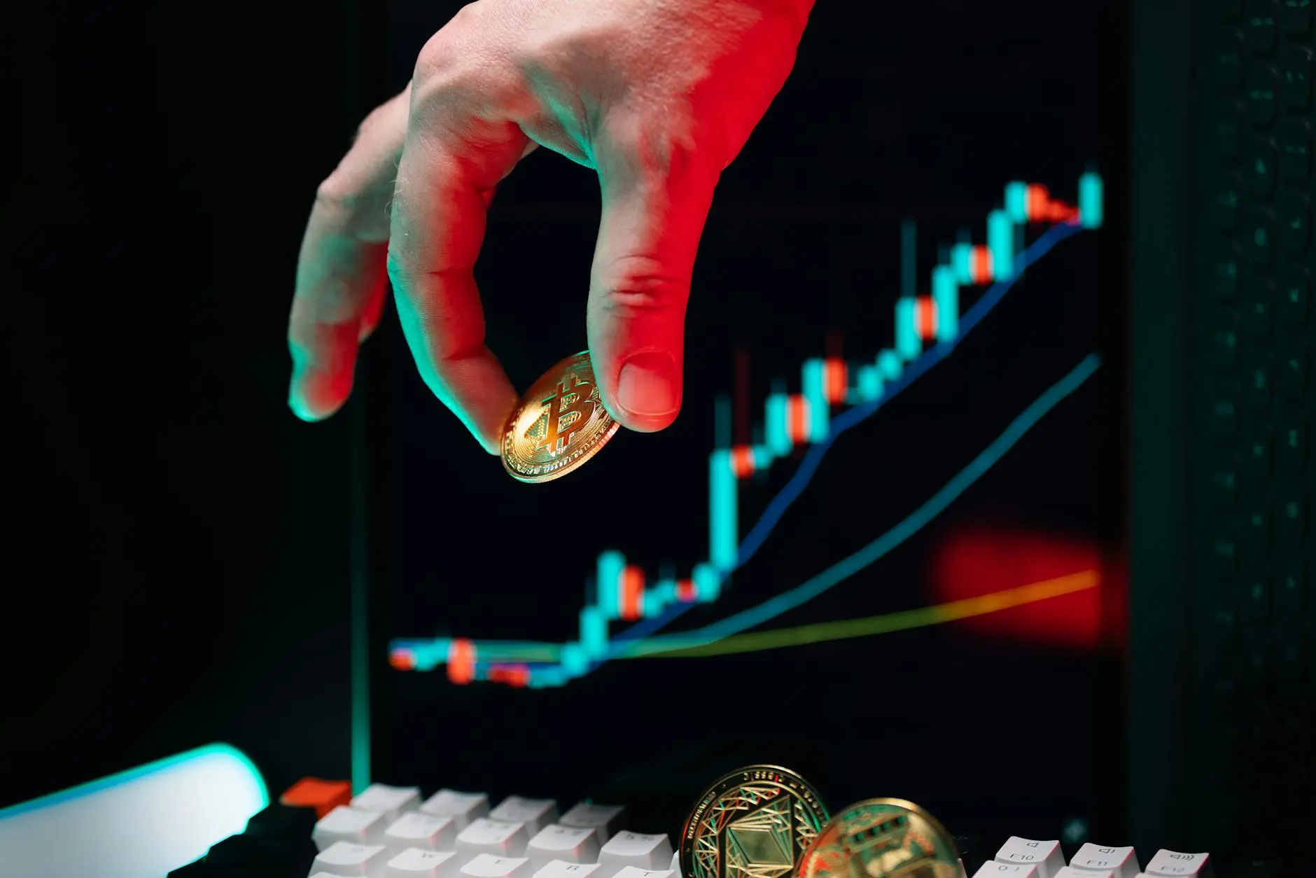 A hand holds a Bitcoin above a keyboard, with a cryptocurrency market chart in the background.