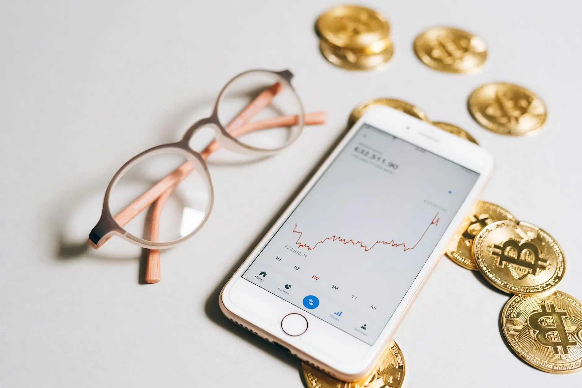 A smartphone displaying a Bitcoin portfolio graph next to glasses and gold Bitcoin coins.