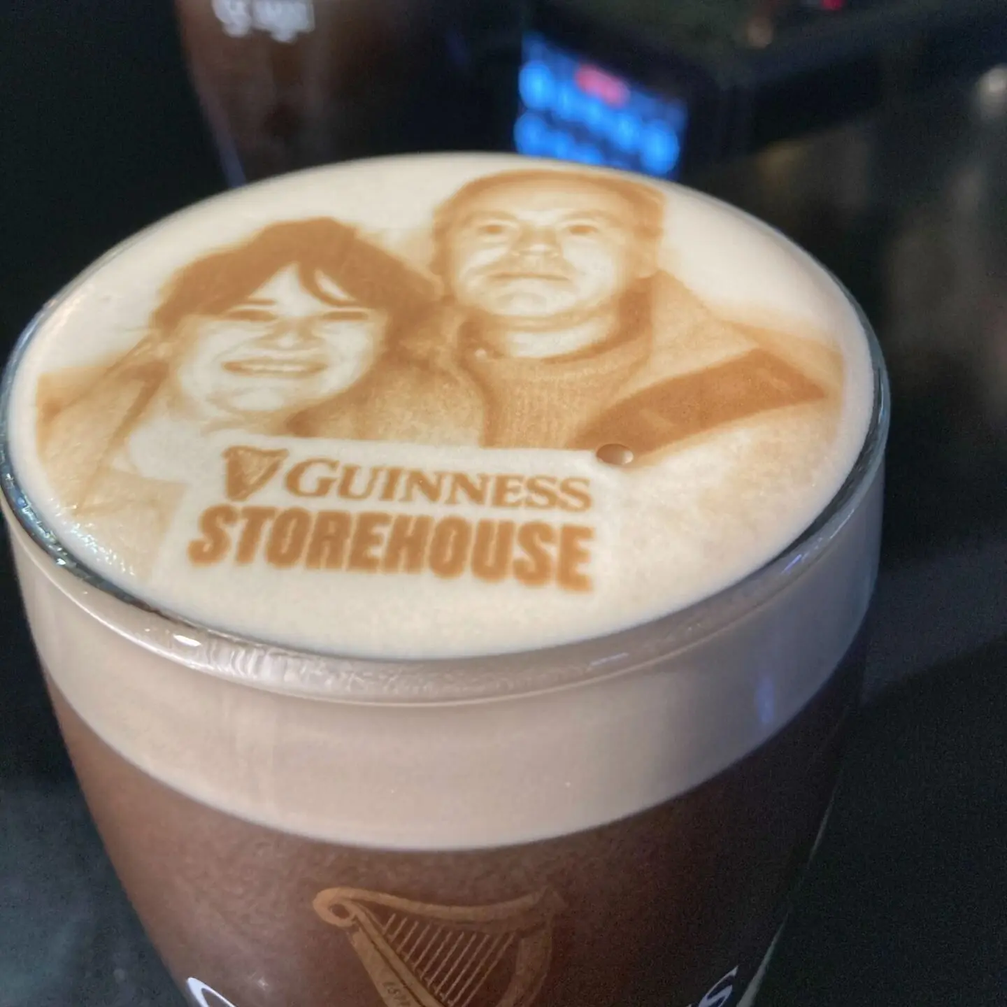 A Guinness pint with a latte art image of two people on the foam, featuring the Guinness Storehouse logo.