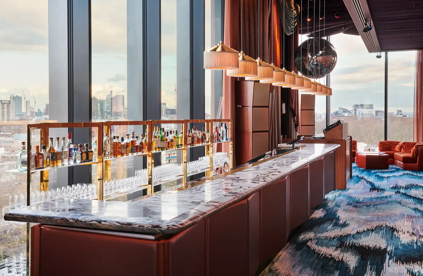Elegant bar interior with marble top, shelves of liquor, and city view through large windows.