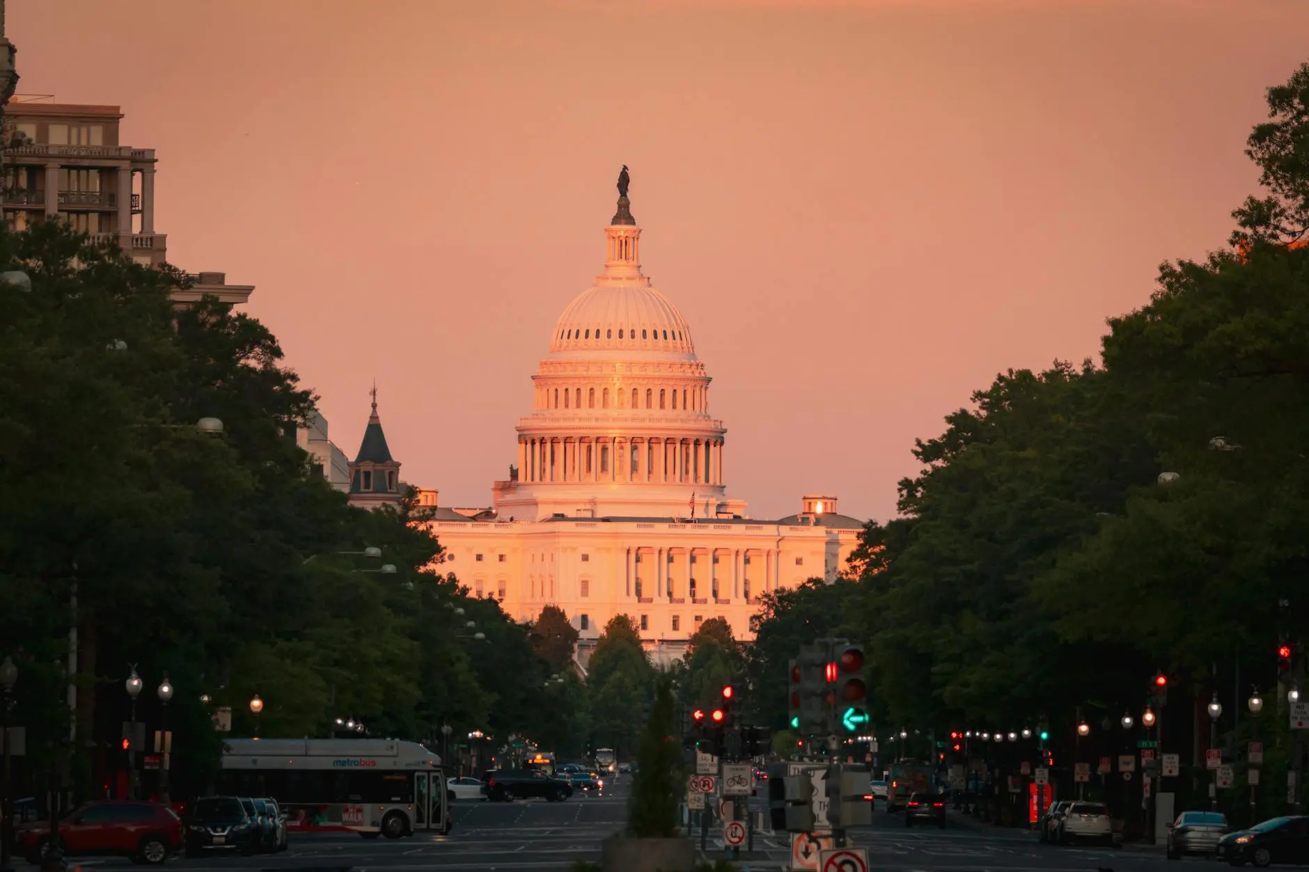 The US Capitol Building lit by sunset, seen from a tree-lined street.