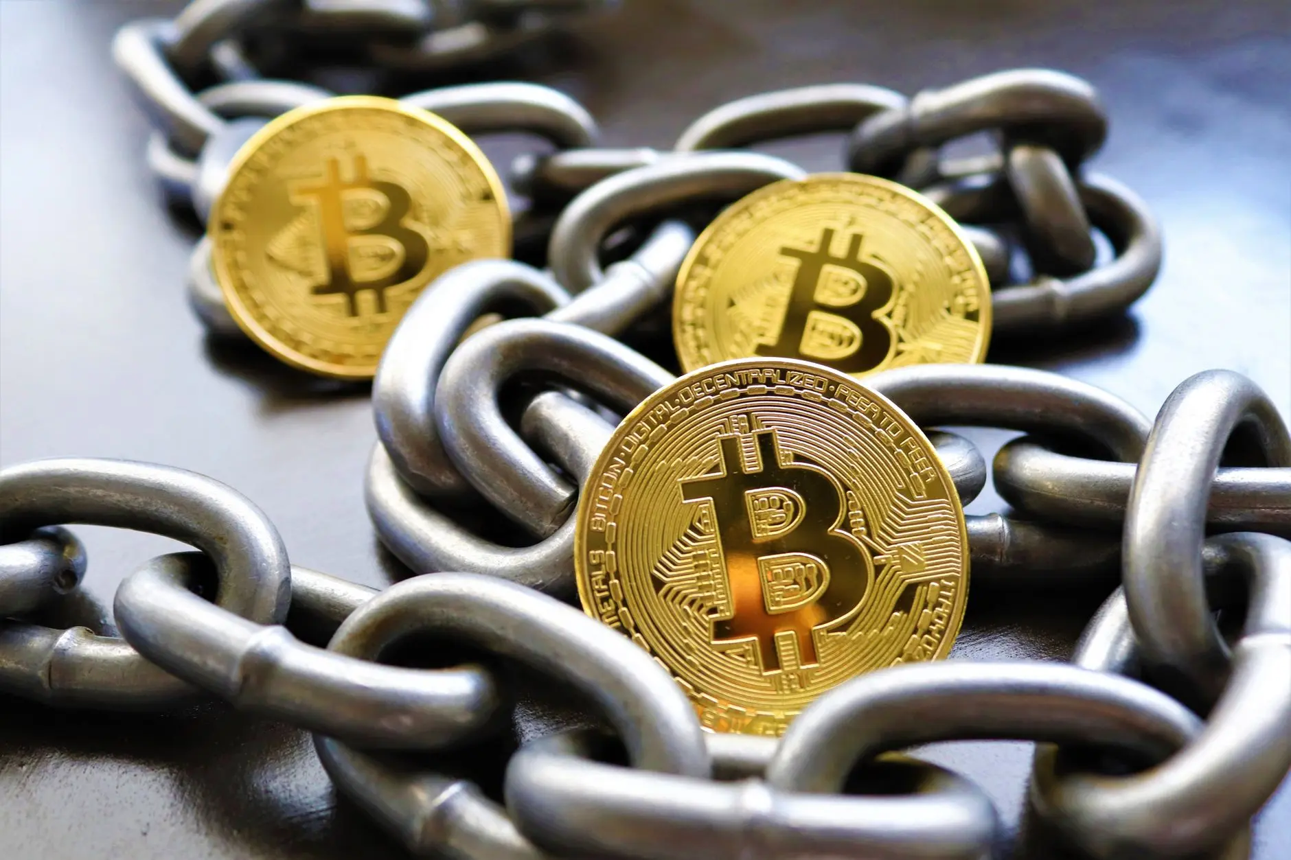 Three gold Bitcoin coins linked by metallic chains on a dark surface.