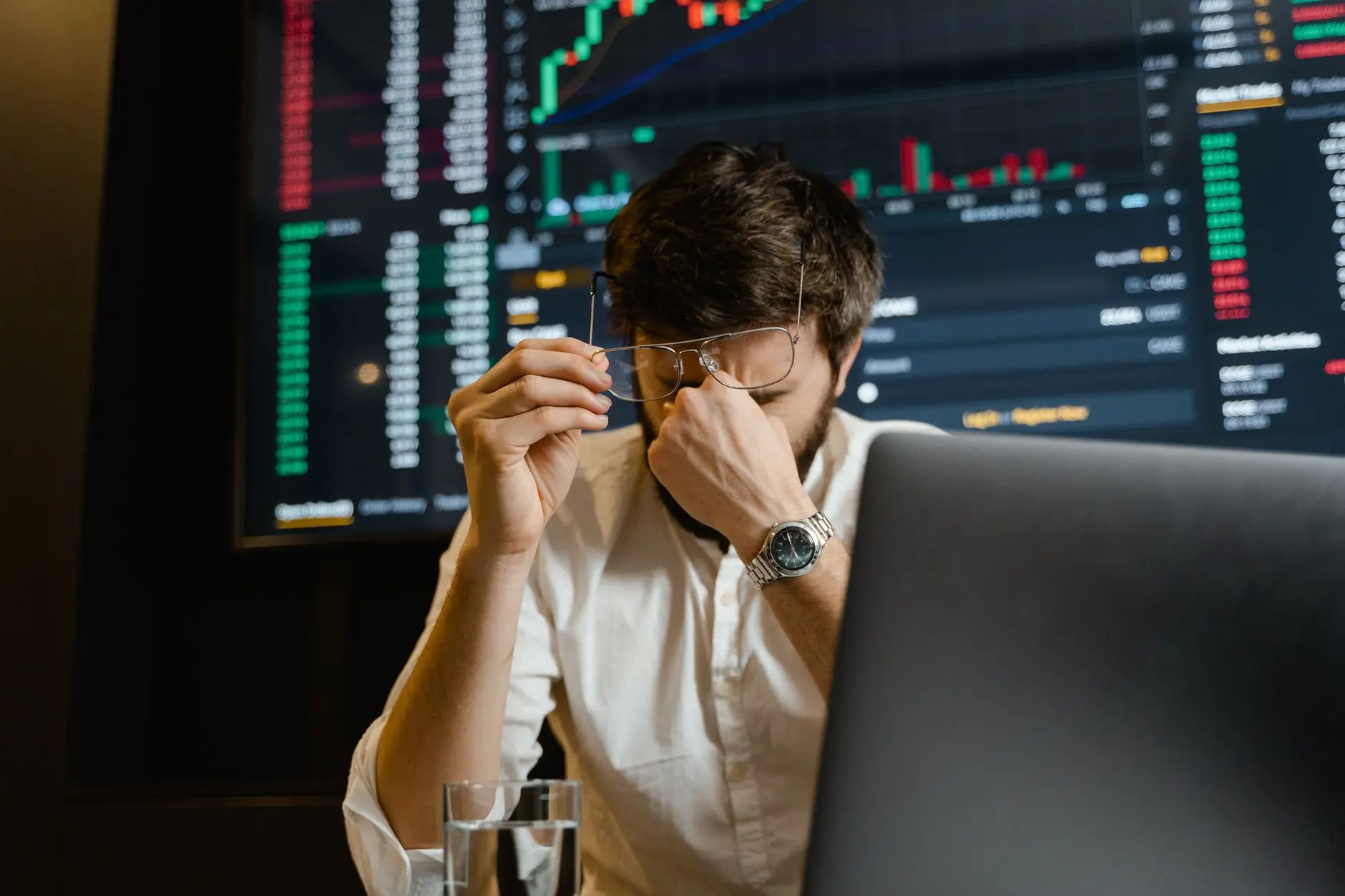 Stressed trader in white shirt examines charts on a dark screen while holding glasses.