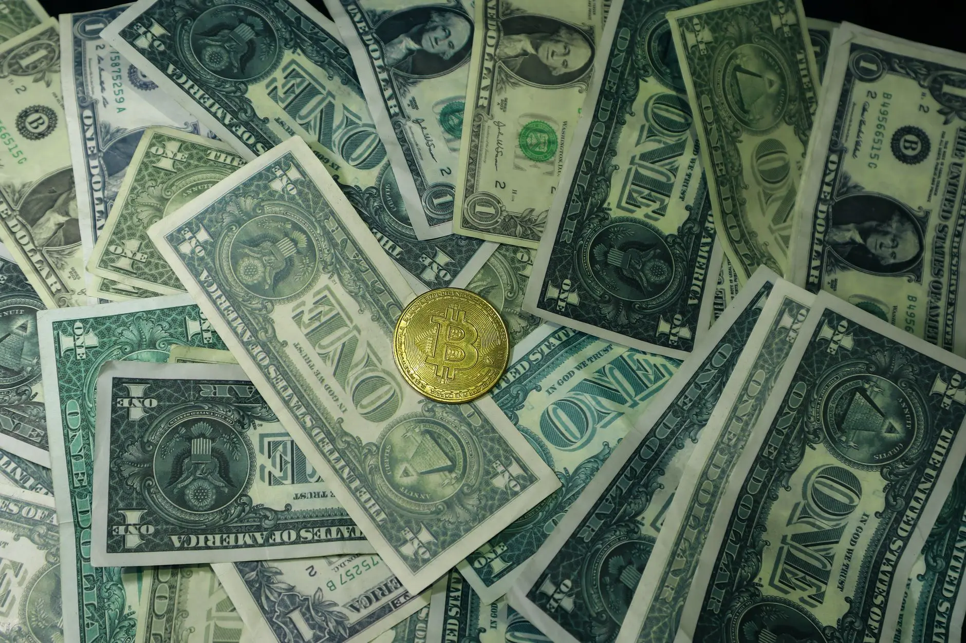 A mix of one dollar bills and a Bitcoin on a dark background.